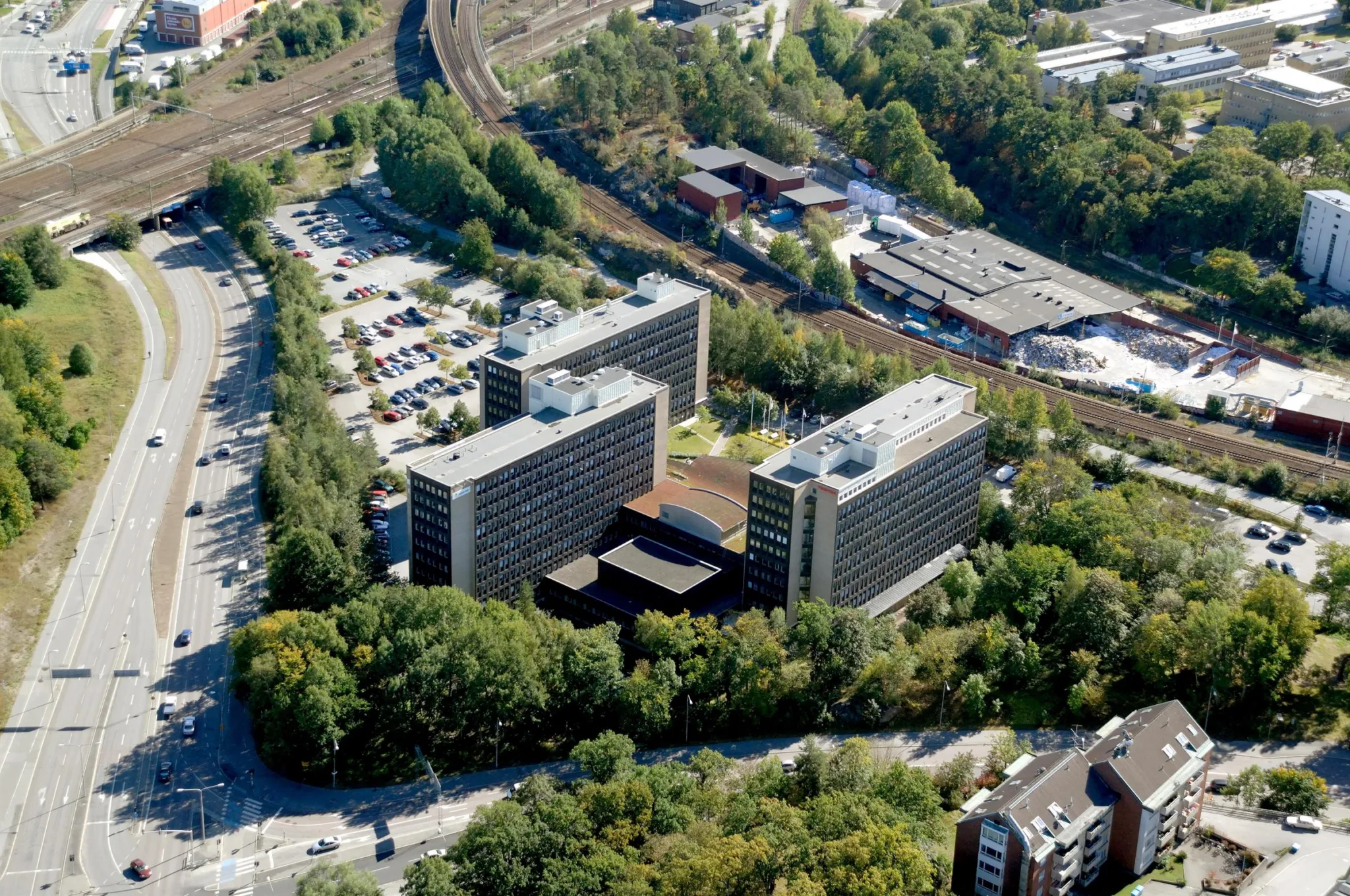 Flygvy över ett område med moderna byggnader omgivna av träd och parkeringsplatser.