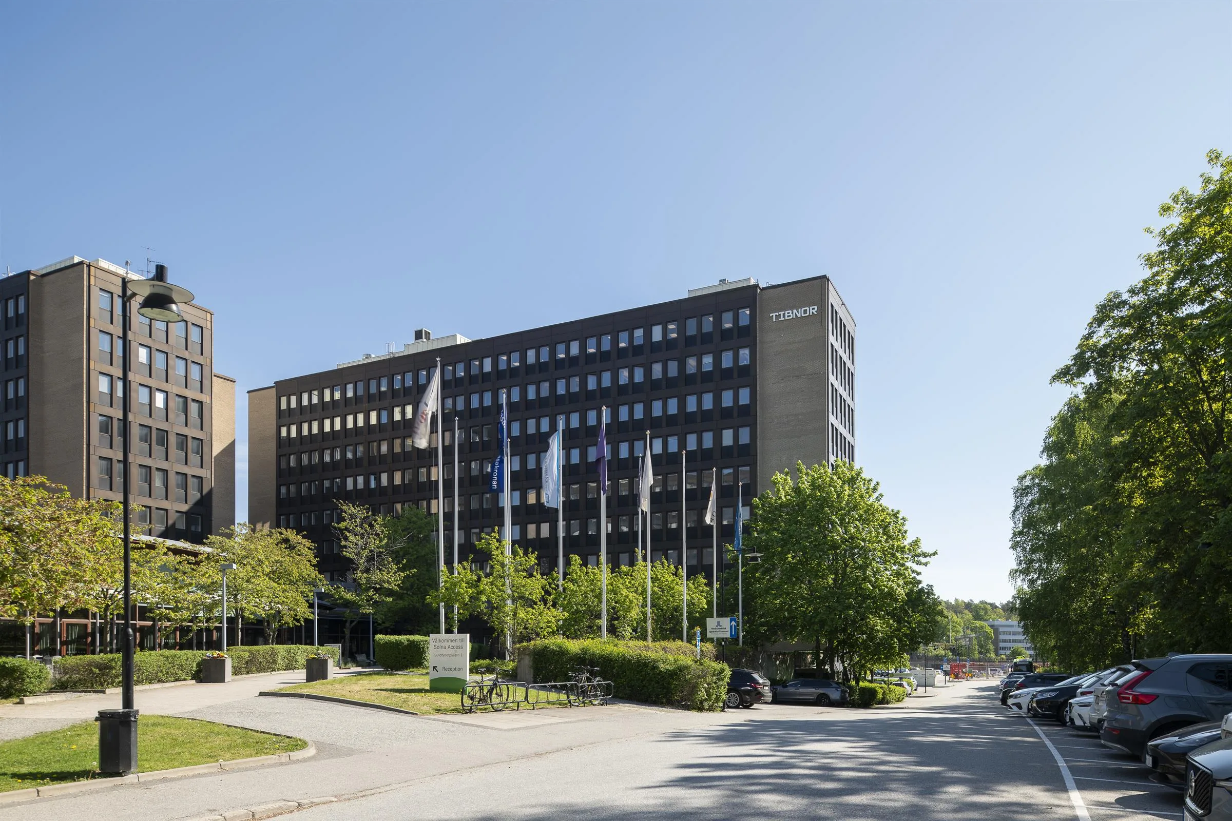 En bred byggnad med flera våningar, flaggor på rad framför och träd i omgivningen.