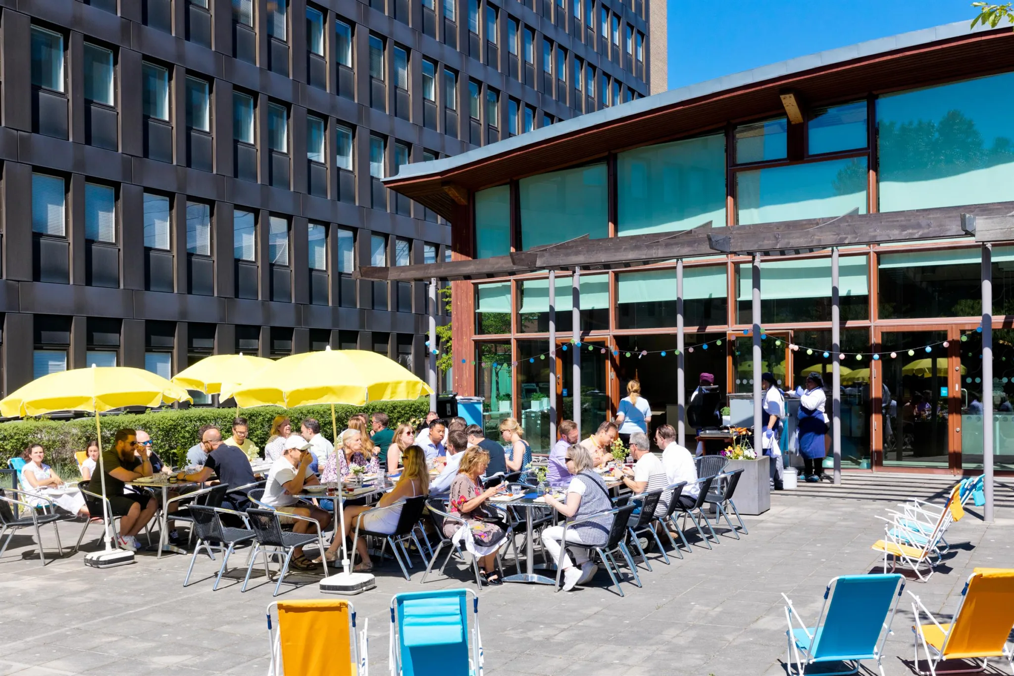 Människor sitter vid bord utomhus under gula parasoller och njuter av mat och dryck; en modern byggnad omges av blå himmel.