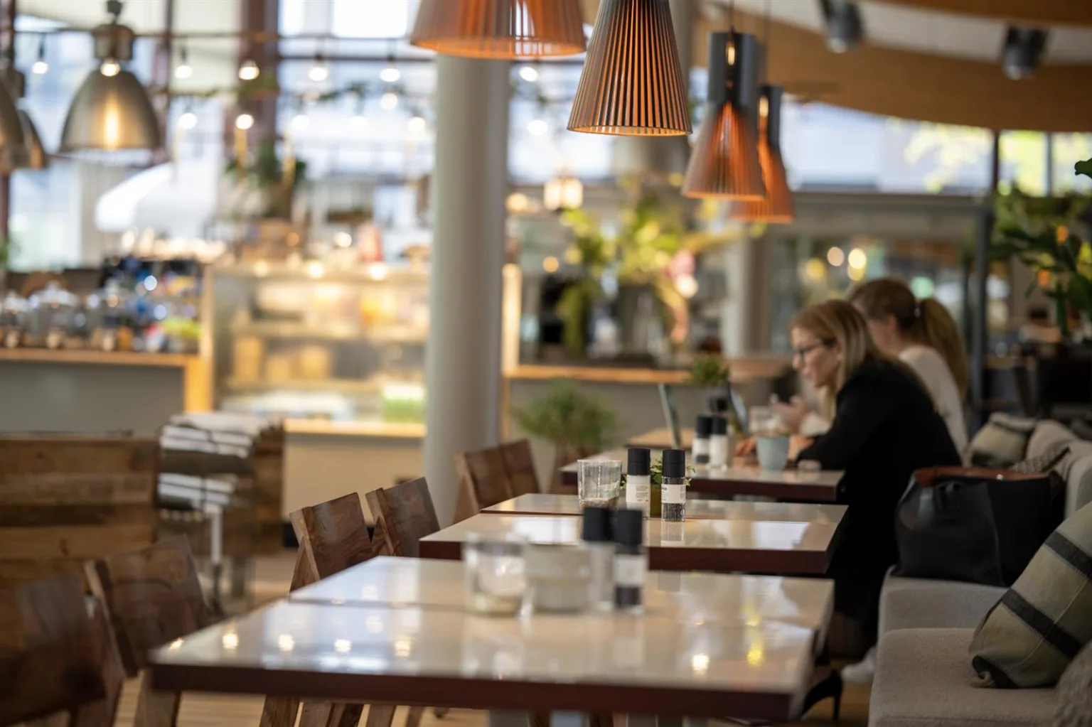 Interiör av en restaurang med bord, stolar och hängande lampor; människor sitter och äter.
