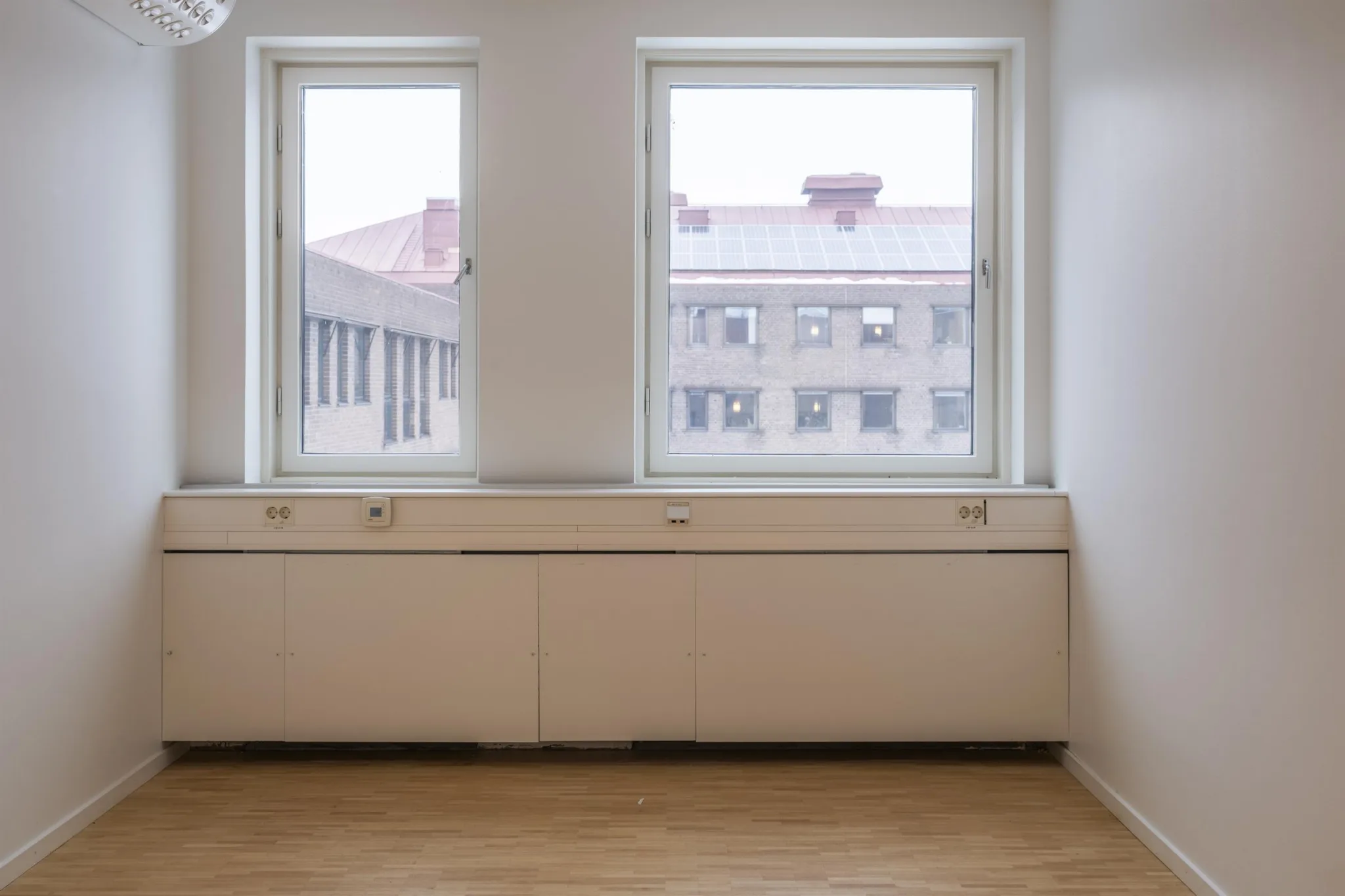 Två fönster med utsikt mot en byggnad, nedanför finns en vit radiator och trägolv.