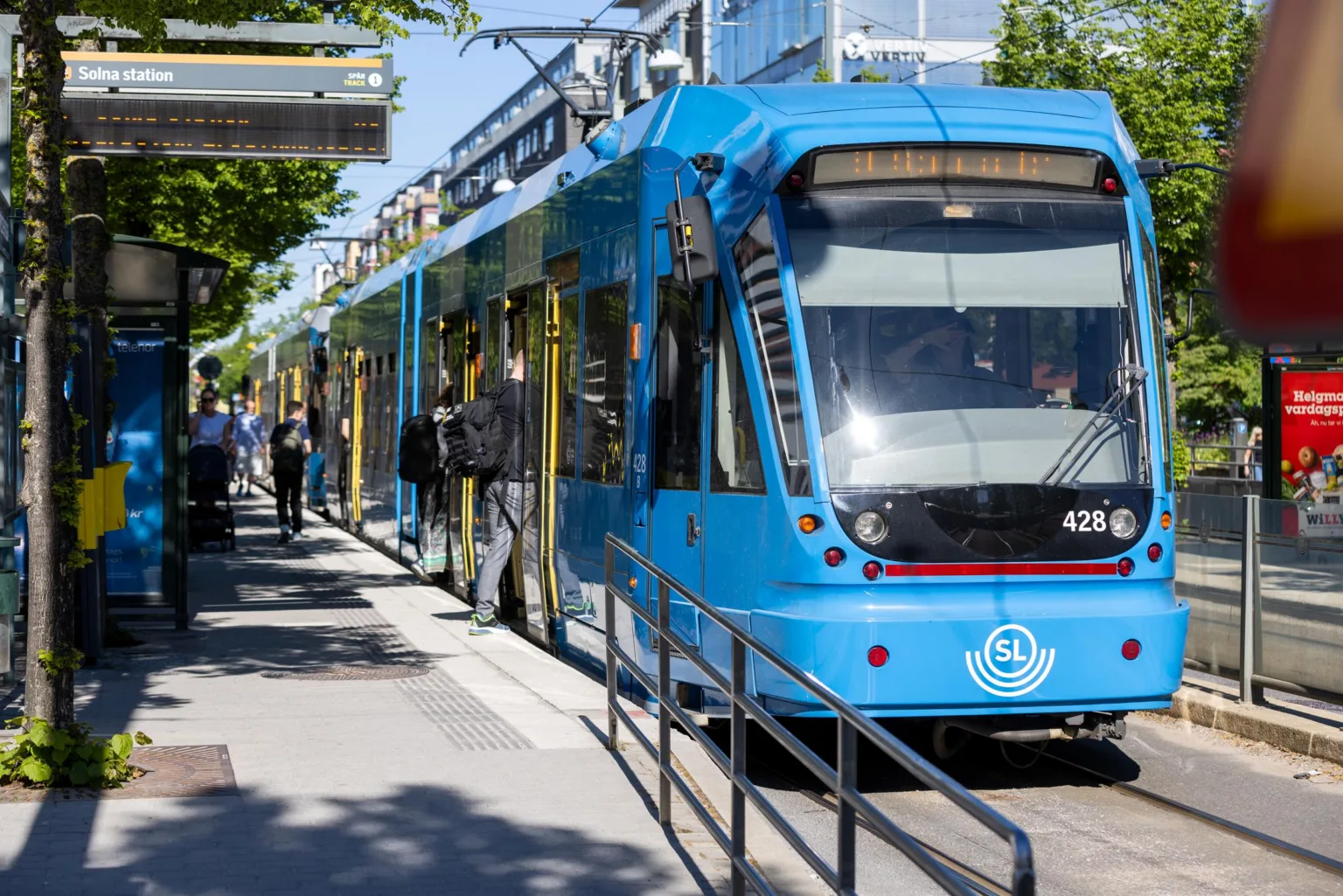 En blå spårvagn står vid en hållplats, där människor går av och på.