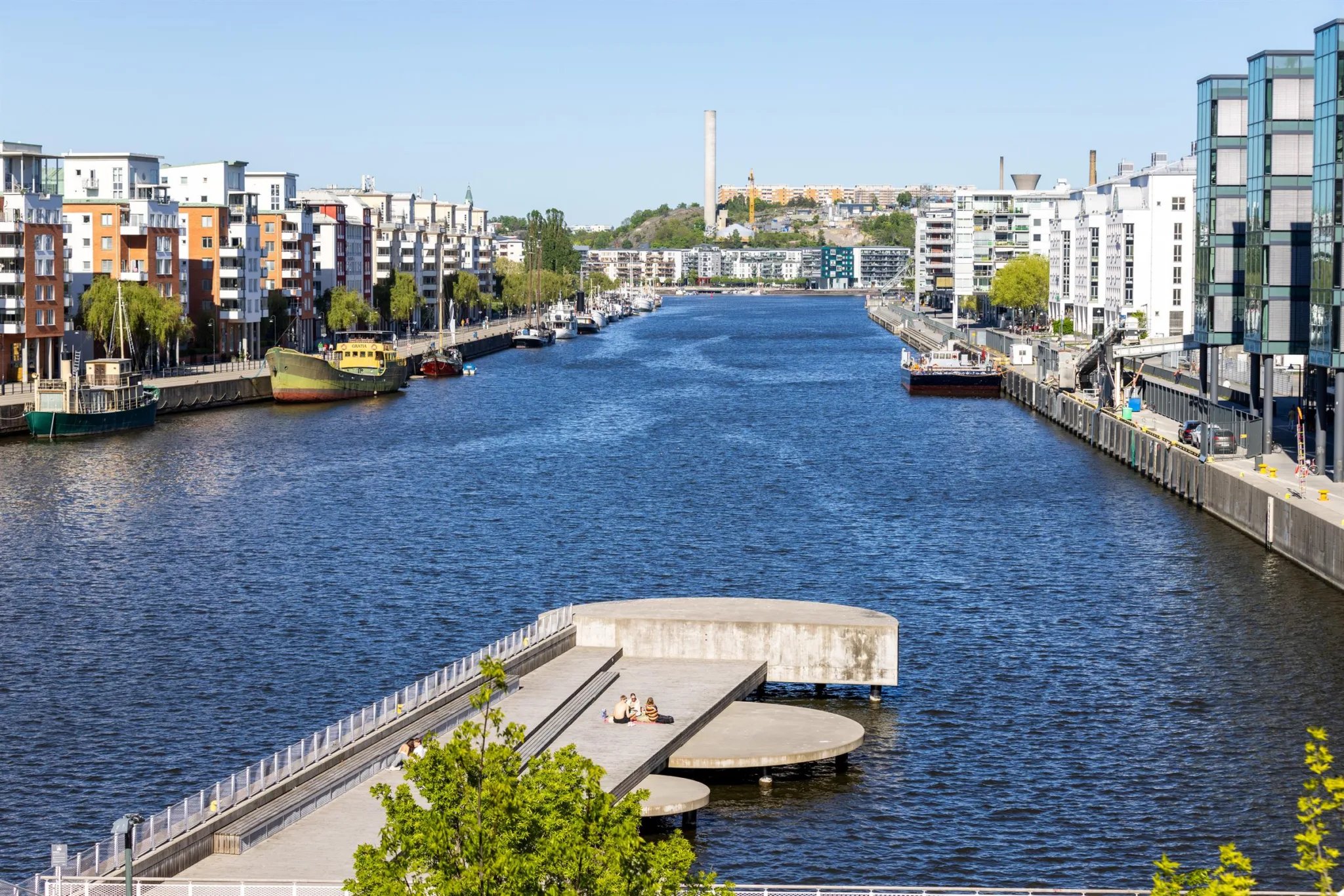 En vattenkanal omgivet av moderna byggnader, småbåtar och en gångbrygga i förgrunden.