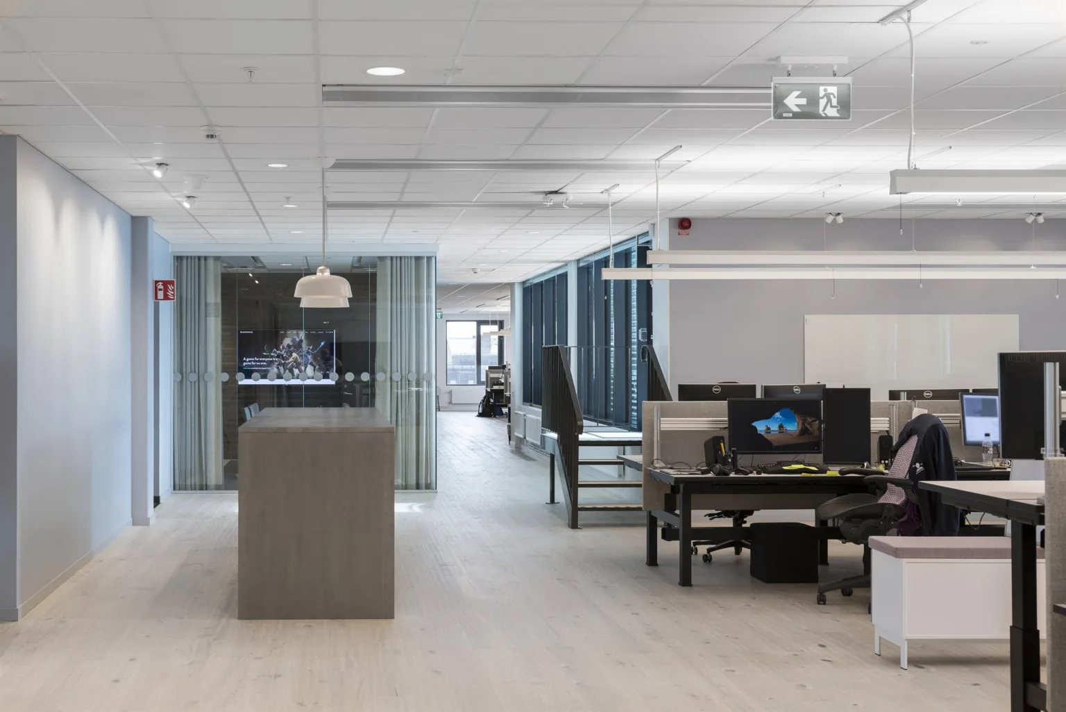 Ett kontor med skrivbord, datorer och glasväggar, med en projektor i taket och ett öppet arbetsutrymme.
