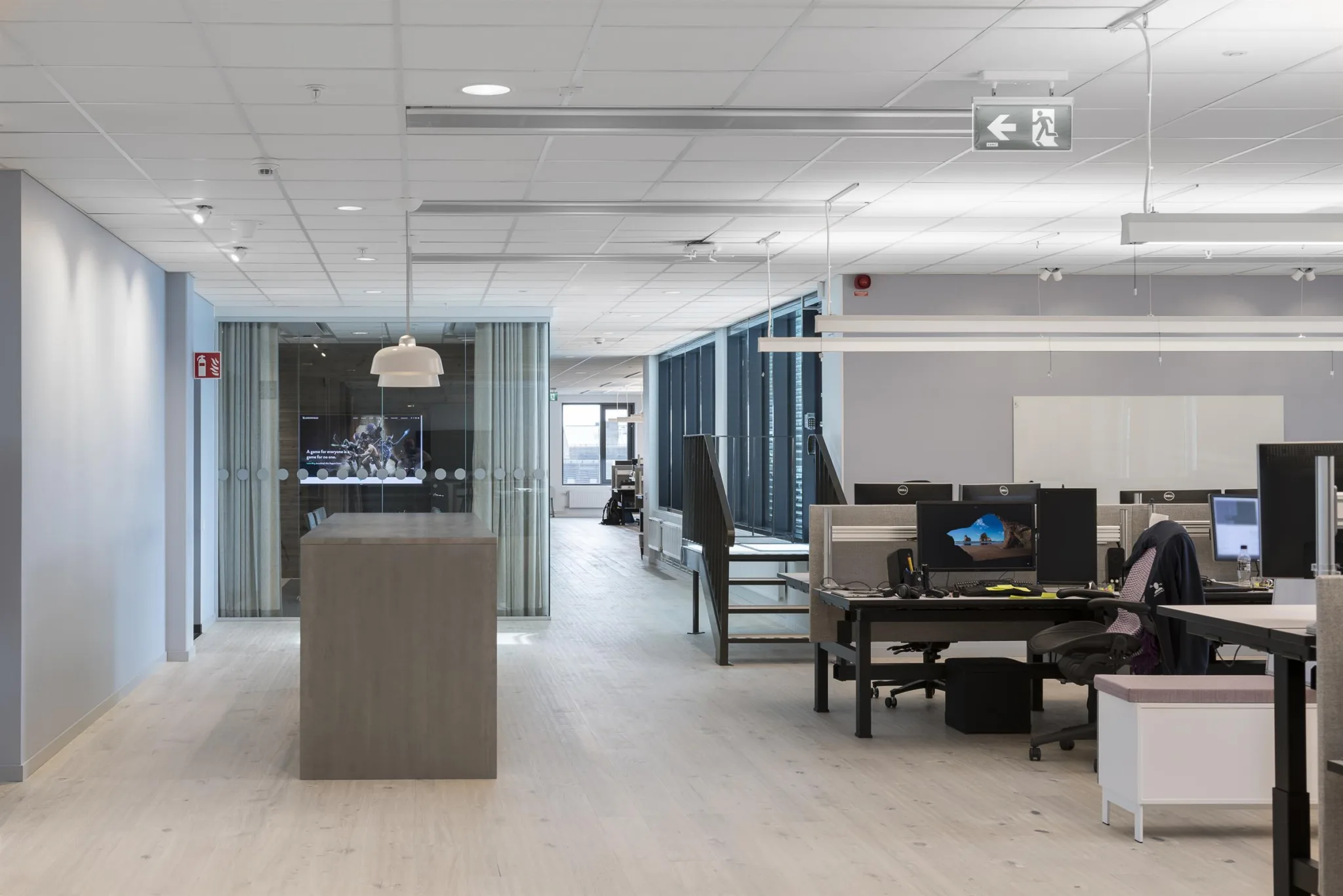 Ett kontor med skrivbord, datorer och glasväggar, med en projektor i taket och ett öppet arbetsutrymme.
