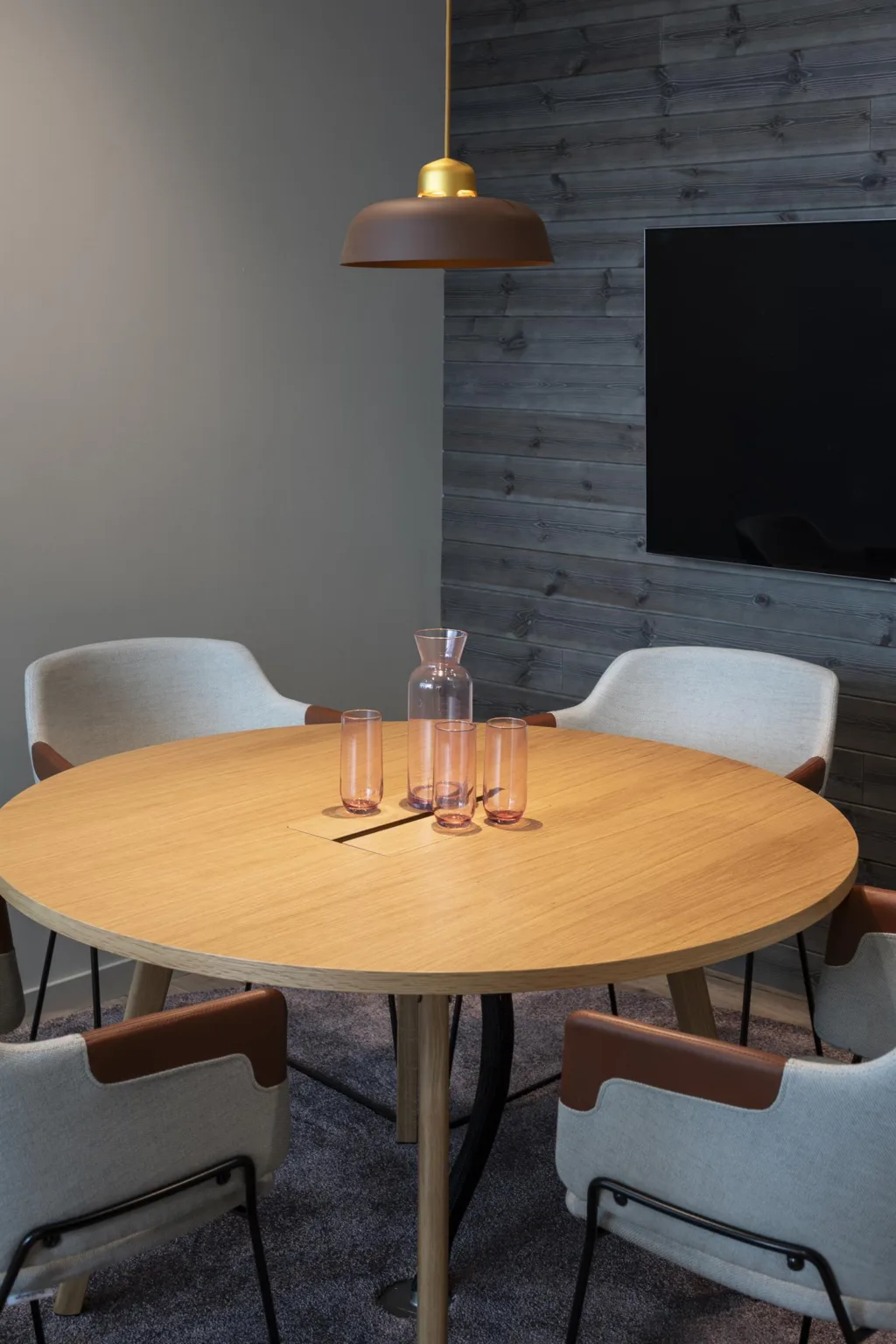 2. Rund träbord omgiven av vita stolar med bruna armstöd, en karaff och glas på bordet, i ett rum med trävägg och TV på väggen.
