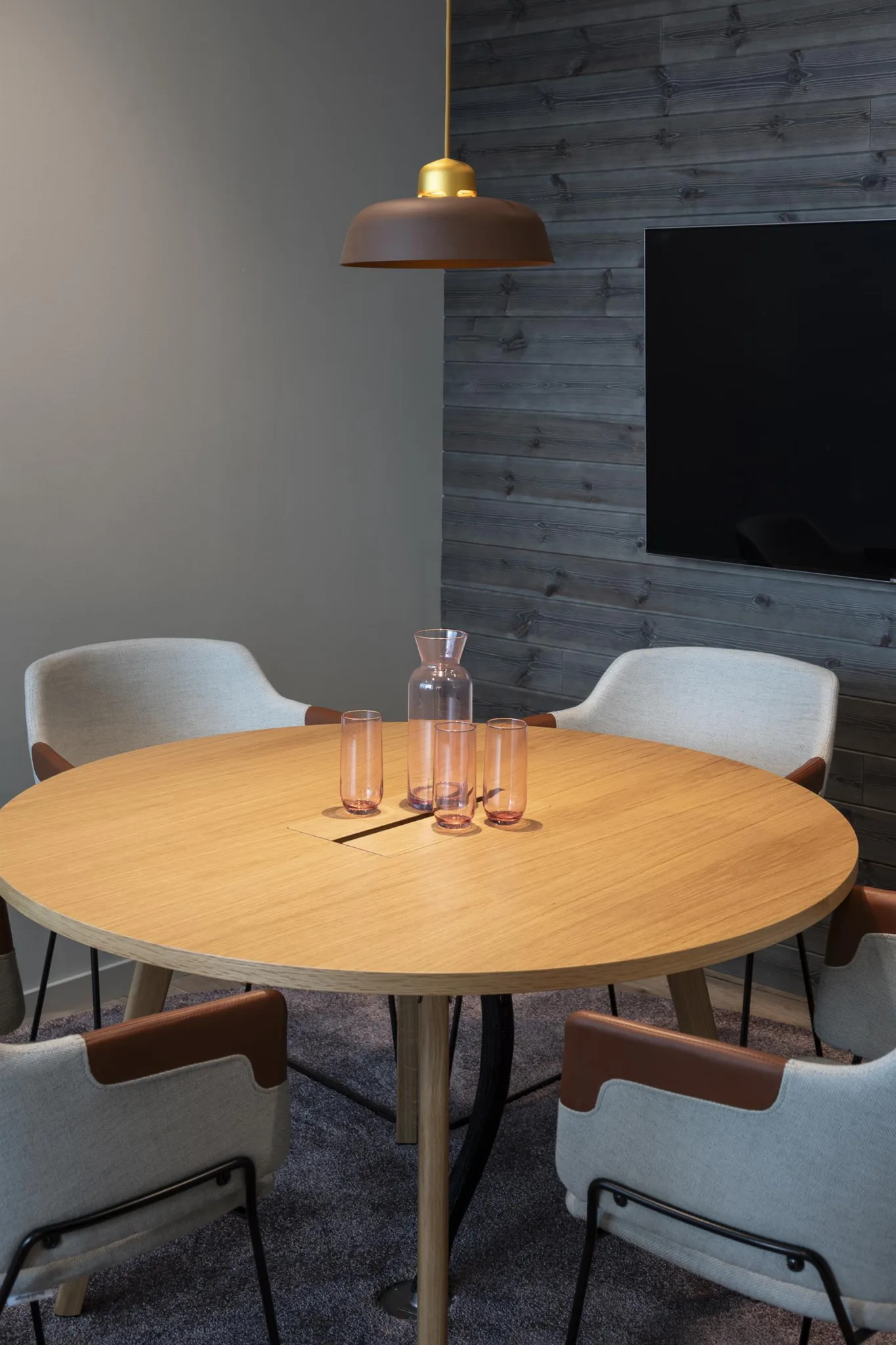 2. Rund träbord omgiven av vita stolar med bruna armstöd, en karaff och glas på bordet, i ett rum med trävägg och TV på väggen.