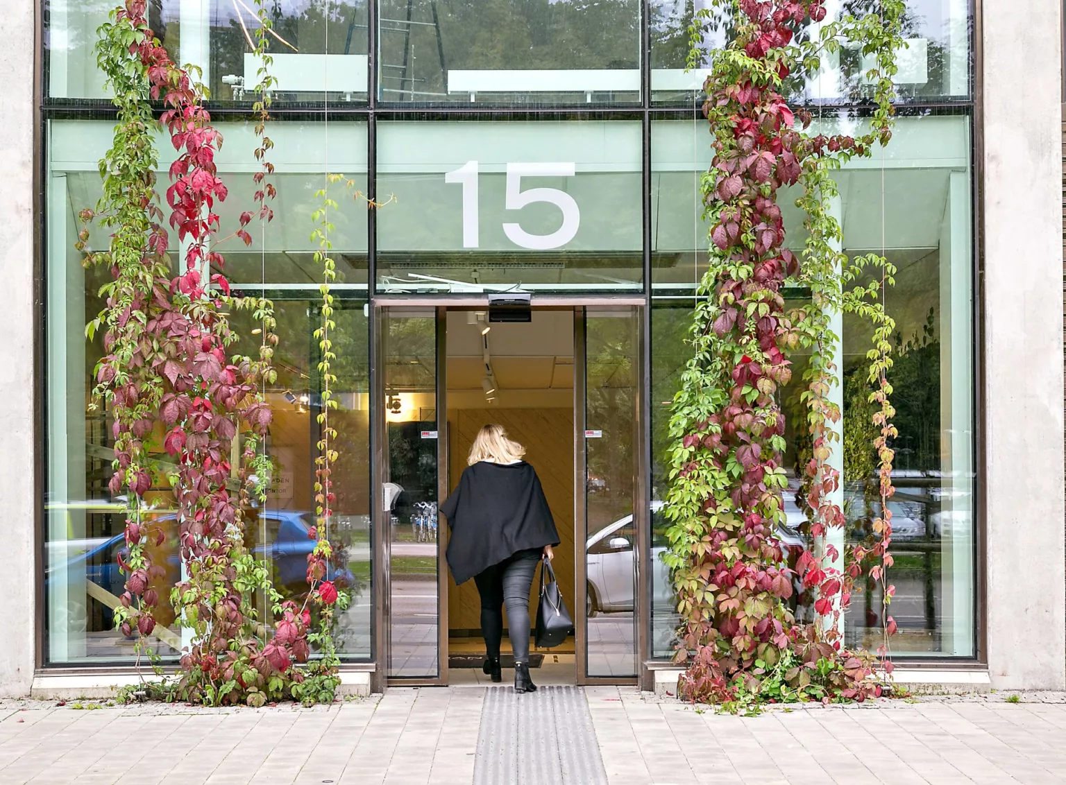 En kvinna klädd i svart går in genom en glasdörr med nummer "15" ovanför, omgiven av klätterväxter med röda och gröna löv.