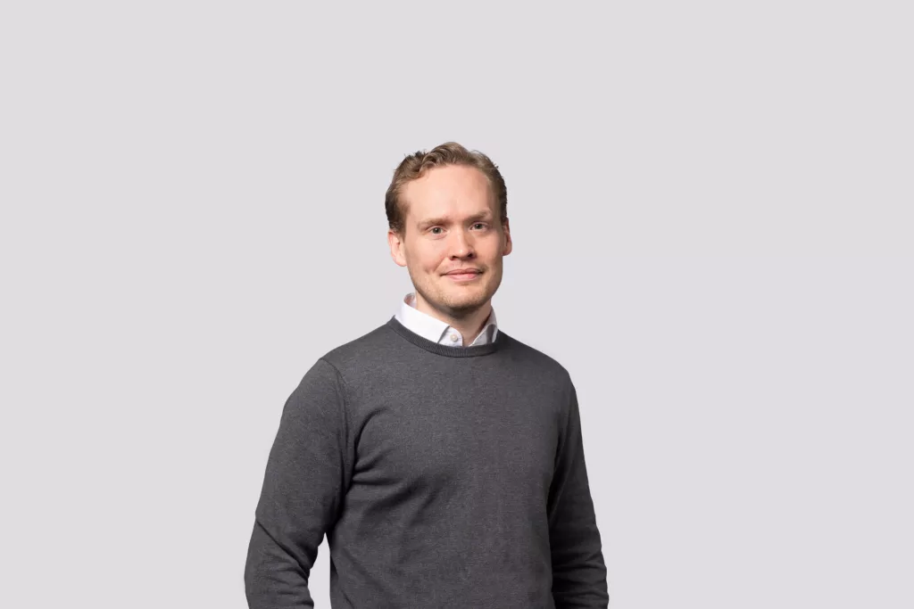 En man med kort ljust hår klädd i en mörkgrå tröja och vit skjorta, står mot en ljusgrå bakgrund.