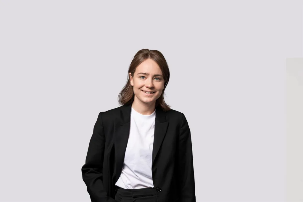 4. En kvinna med axellångt hår, iklädd en svart kavaj och vit topp, ler och står framför en ljus bakgrund.