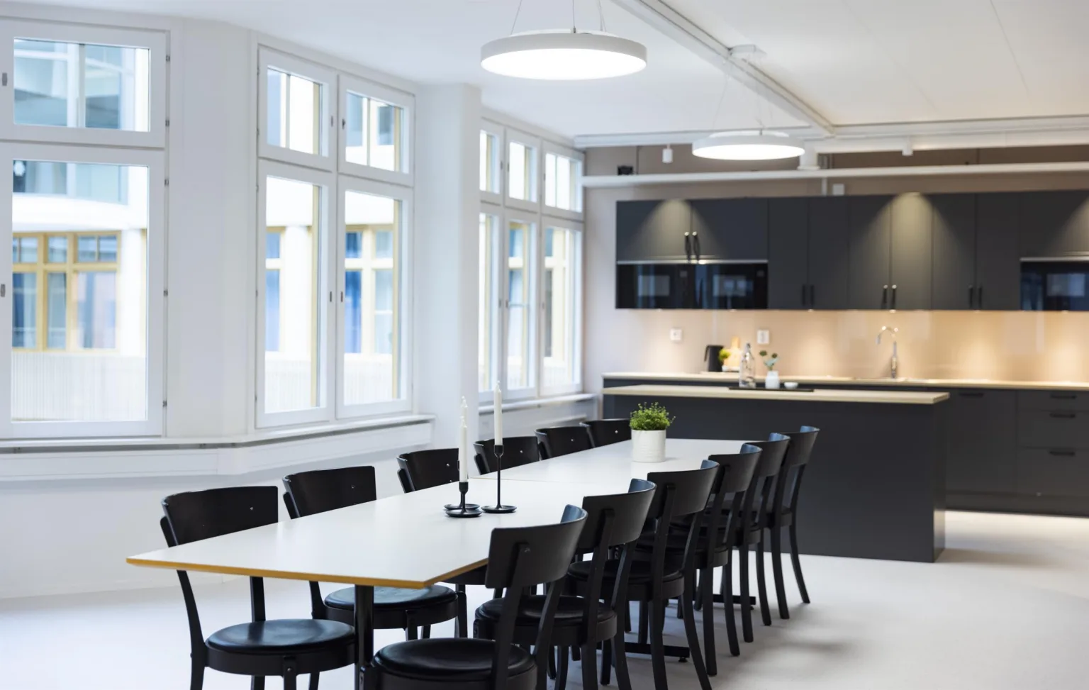 Ett modernt konferensrum med lång vit bordsskiva, svarta stolar och ett kök i bakgrunden.