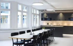 Ett modernt konferensrum med lång vit bordsskiva, svarta stolar och ett kök i bakgrunden.