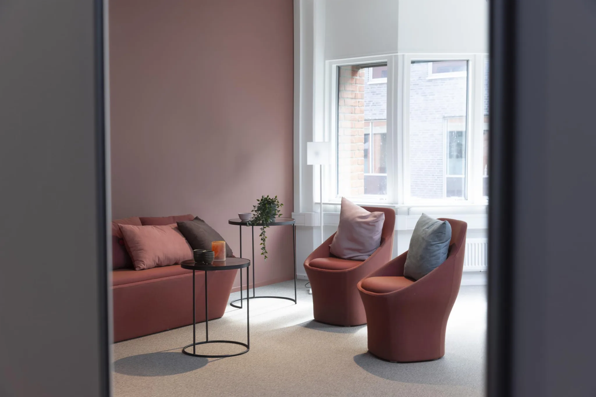 Ett stilrent loungeområde med rosa fåtöljer, en soffa, små bord och stora fönster som släpper in naturligt ljus.