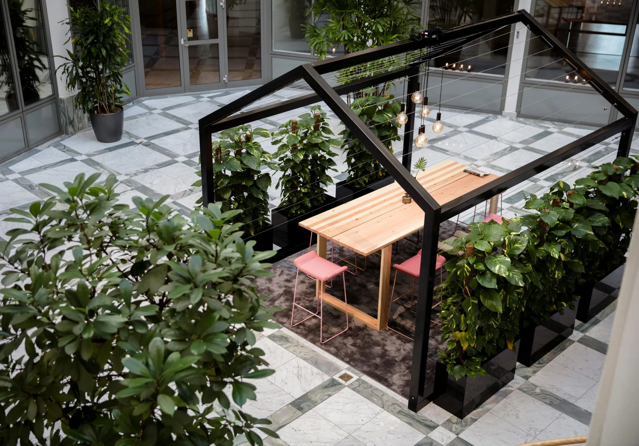 5. Ett uterum med ett träbord och rosa stolar under en svart metallstruktur, omgiven av grönskande växter.