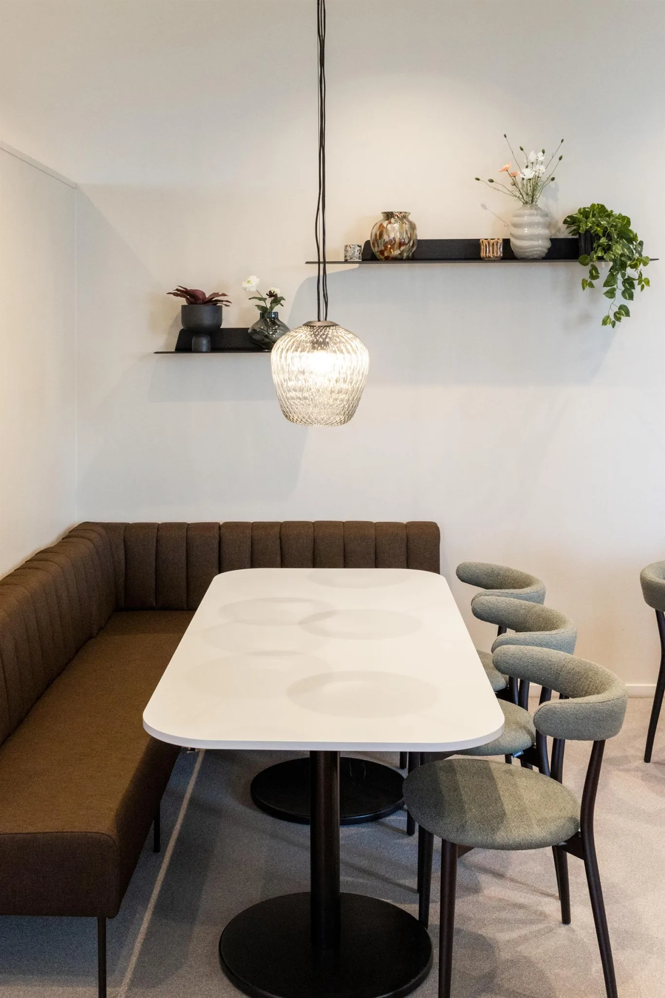 Ett modernt matbord med en brun hörnsoffa, fyra gråa barstolar och en hängande lampa ovanför, med hyllor och växter i bakgrunden.