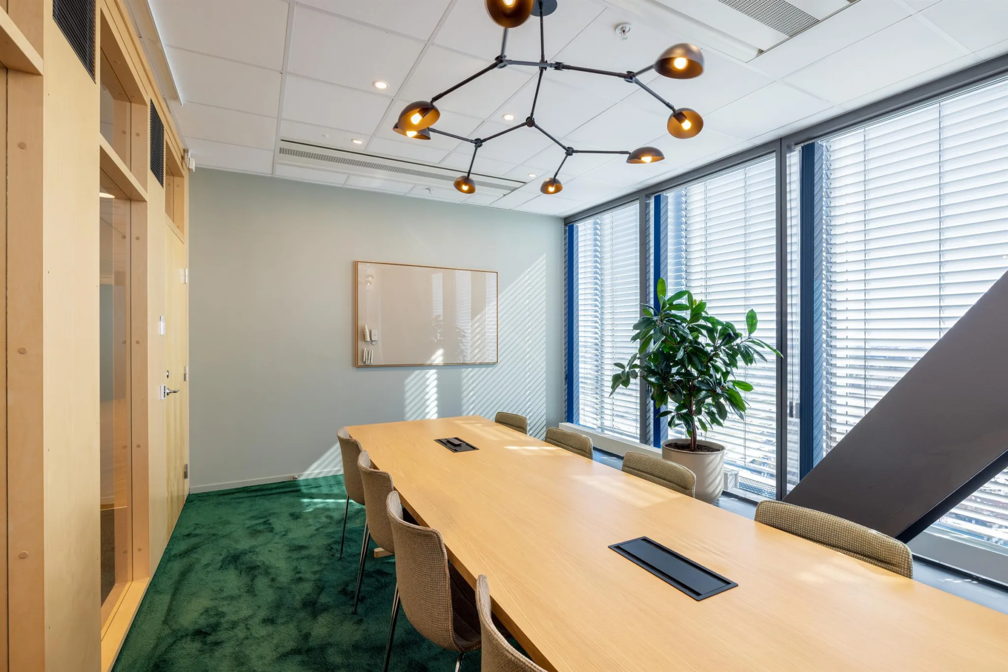 5. Ett konferensrum med ett långt bord, stolar, en whiteboard, en stor växt och stora fönster som släpper in dagsljus.
