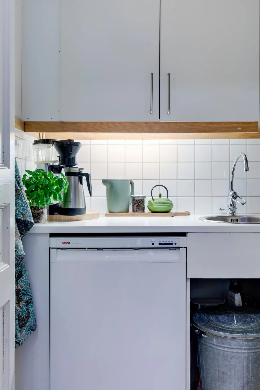 Ett litet kök med vit inredning, diskbänk, kaffebryggare och grön växt.
