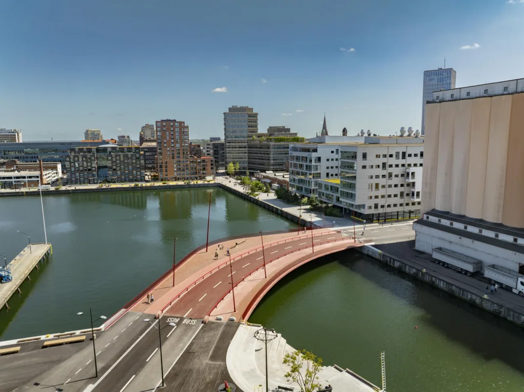 En röd gångbro sträcker sig över en kanal, omgiven av byggnader och stadsmiljö.