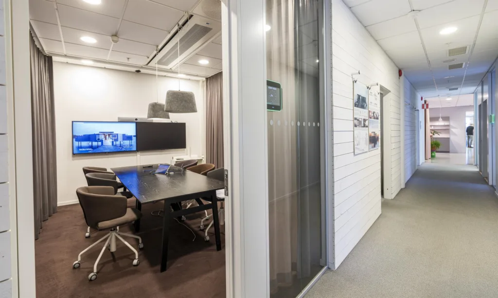4. Ett konferensrum med ett mörkt bord, bruna stolar och en stor skärm på väggen, synligt genom en glasvägg.