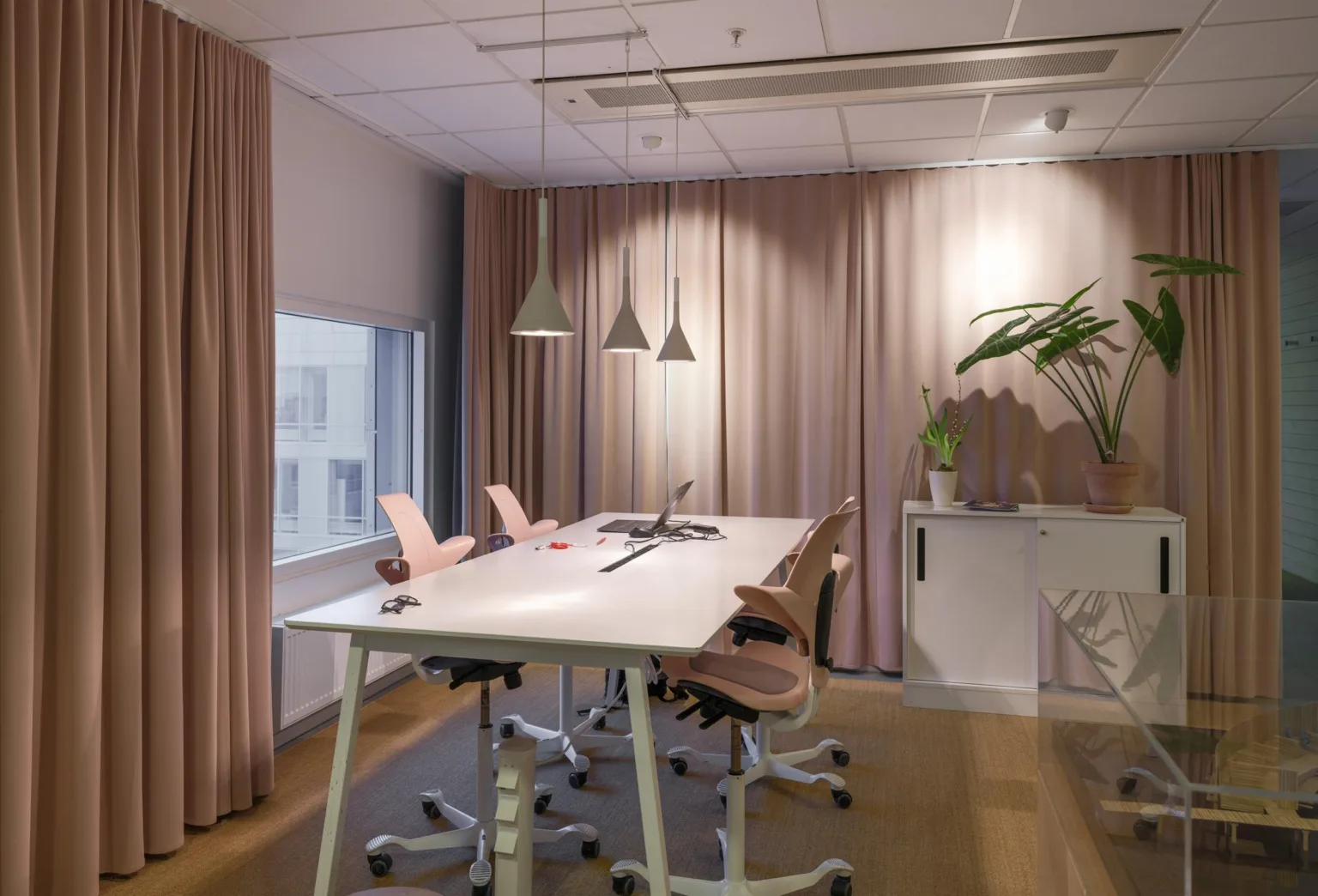 Ett arbetsrum med ett långt vitt bord, ergonomiska stolar och hängande lampor, samt gardiner och en växt i bakgrunden.