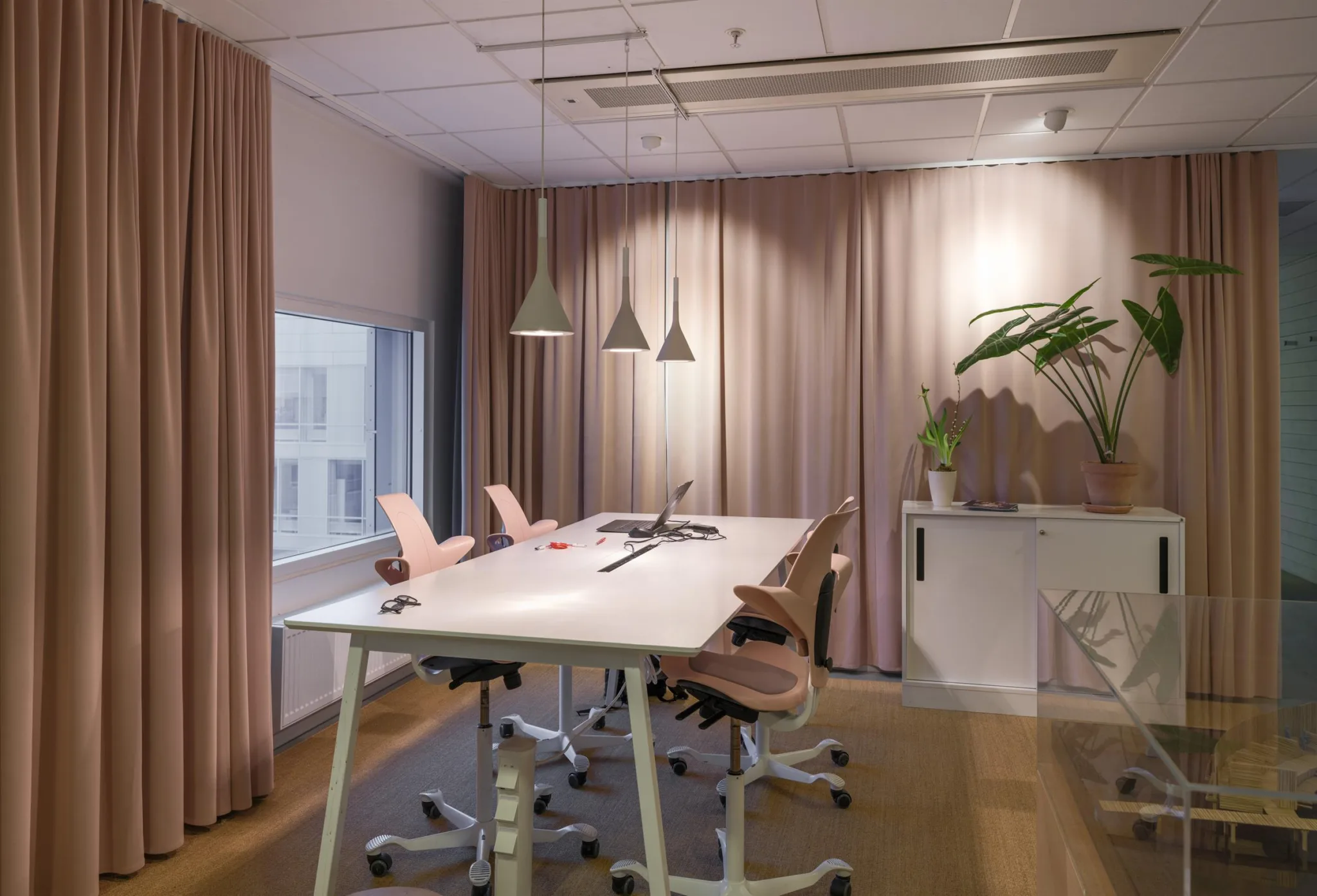 Ett arbetsrum med ett långt vitt bord, ergonomiska stolar och hängande lampor, samt gardiner och en växt i bakgrunden.
