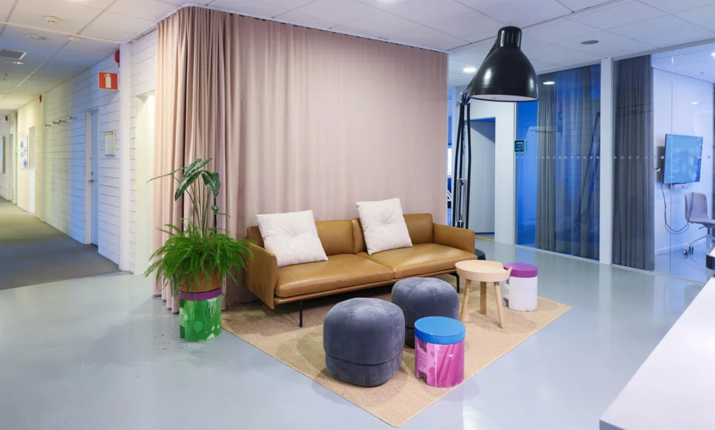 1. Ett väntrum med en brun soffa, två vita kuddar, en grön växt och färgglada sittpuffar på en matta under en stor svart lampa.