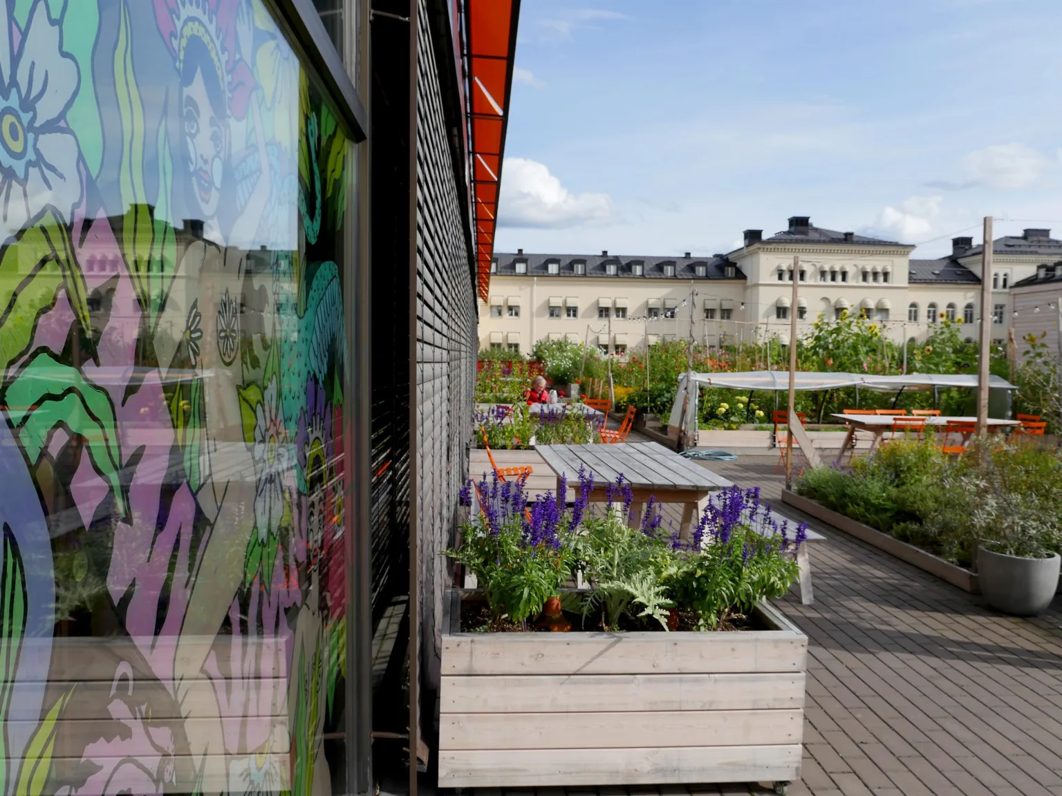 1. En utomhusterrass med planteringslådor fyllda med blommor och gröna växter, samt ett bostadshus i bakgrunden.
