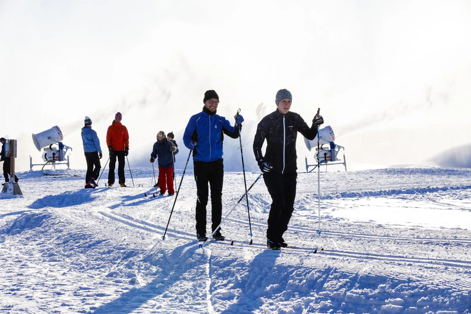 Två personer åker längdskidor på en snötäckt slätt med fler skidåkare i bakgrunden.
