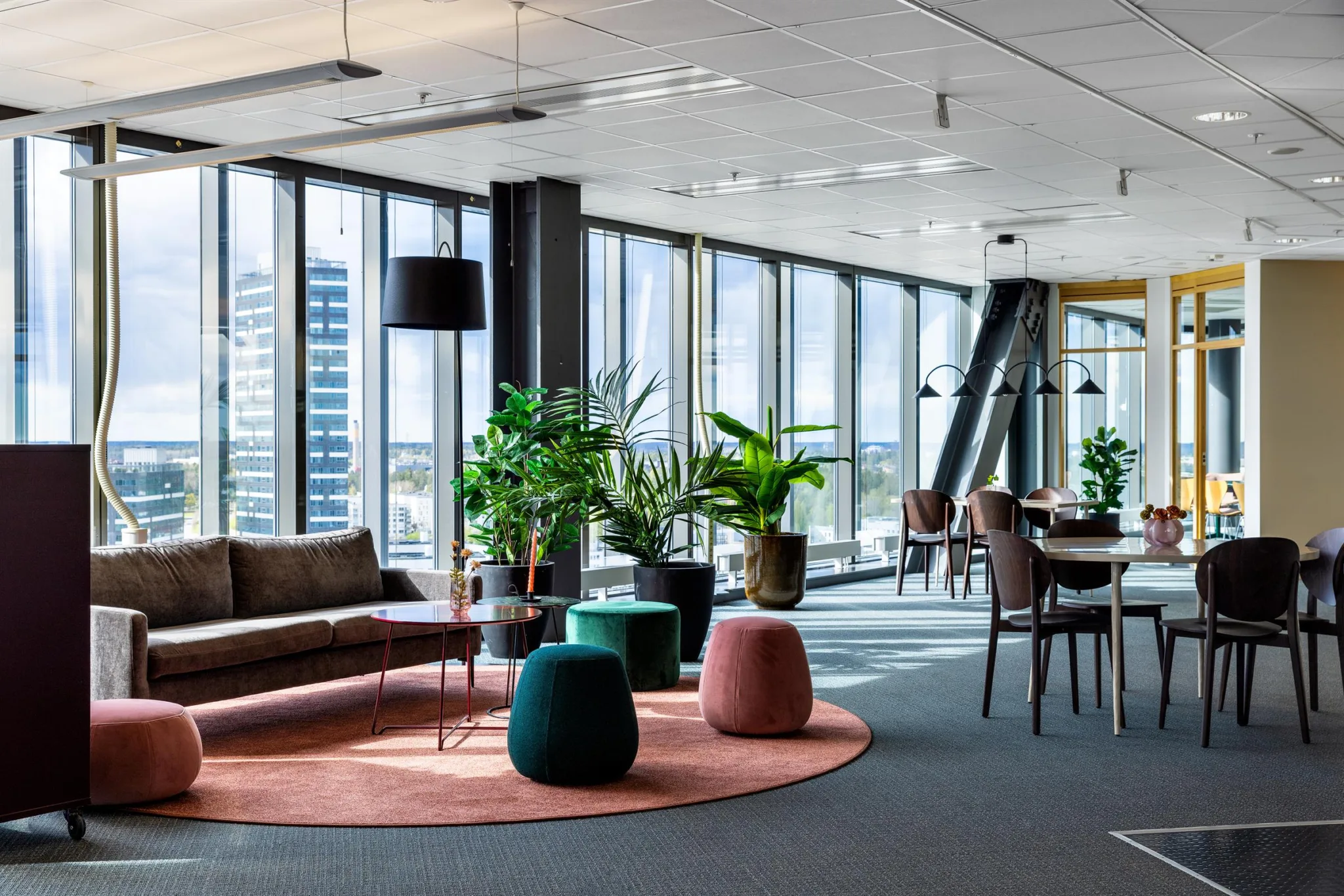 En modern lounge med soffa, sittpuffar, gröna växter och matbord nära stora glasfönster med utsikt.