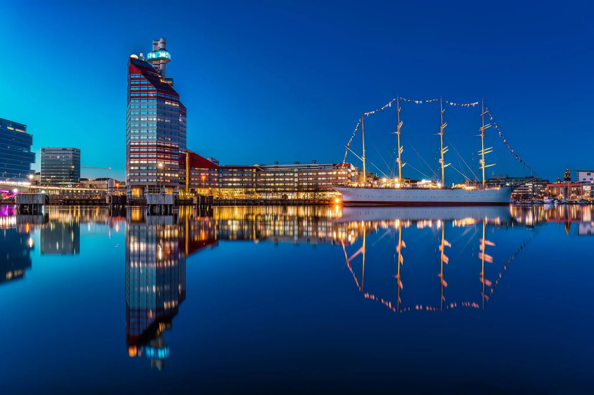 2. En nattlig vy över Göteborgs hamn med Lilla Bommen-byggnaden och ljusdekorerade båtar som speglas i vattnet.