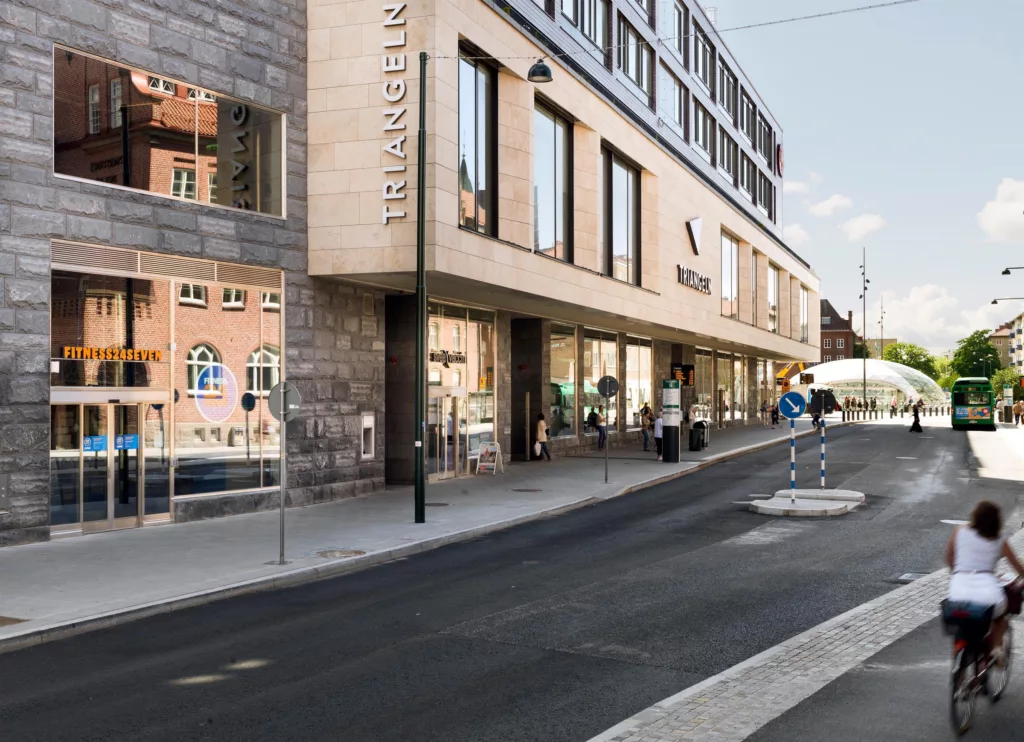Gatubild av byggnaden "Triangeln", med glasfasad och butikeringångar längs trottoaren.