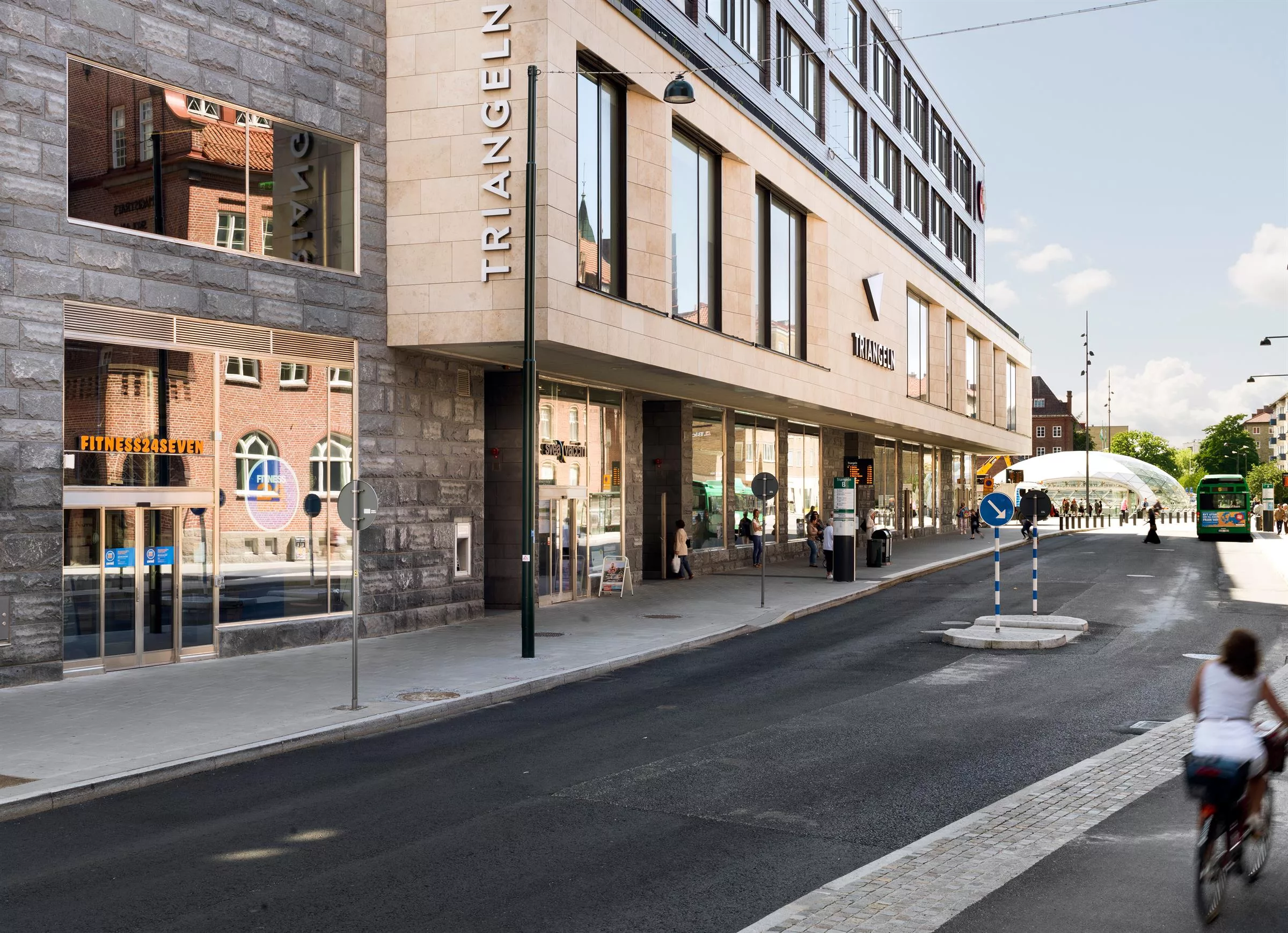 Gatubild av byggnaden "Triangeln", med glasfasad och butikeringångar längs trottoaren.
