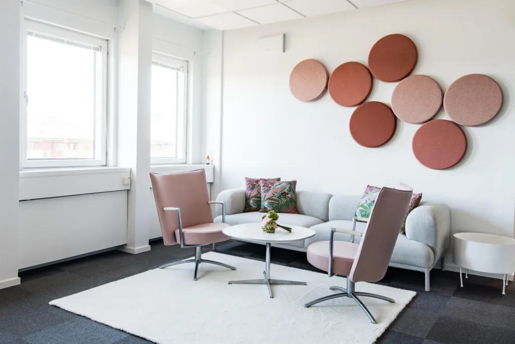 Ett ljust vardagsrum med två rosa stolar, en vit soffa, ett litet vitt bord och runda orangea dekorationer på väggen.