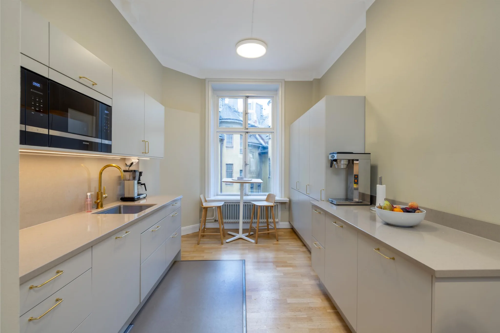 Ett modernt kök med ljusa skåp, bänkskivor, mikrovågsugn och kaffemaskin, samt ett stort fönster.