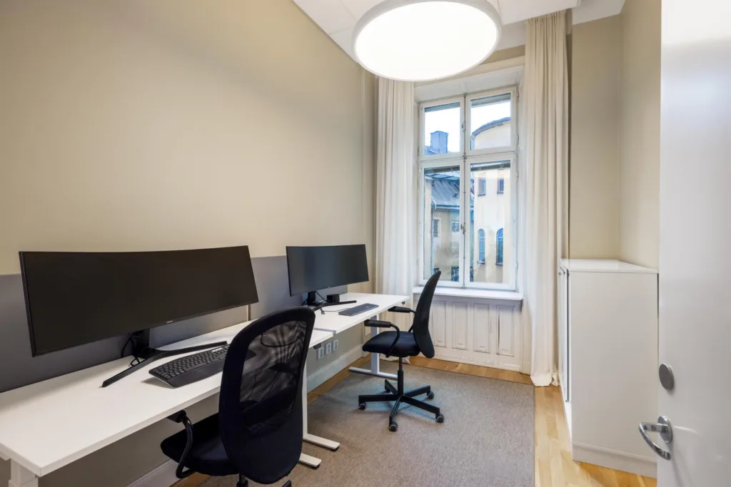 Ett mindre kontorsrum med två skrivbord, kontorsstolar och datorskärmar, nära ett fönster med utsikt utåt.