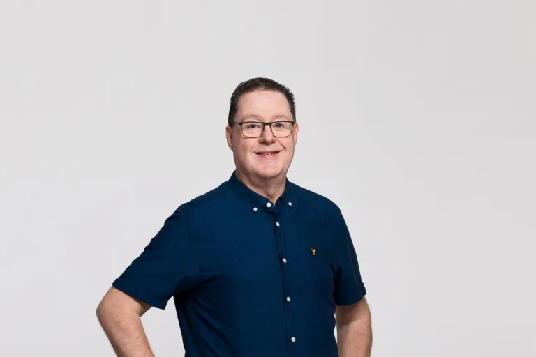 En man med kort hår och glasögon, klädd i mörkblå skjorta, står med ena handen på höften mot en ljus bakgrund.