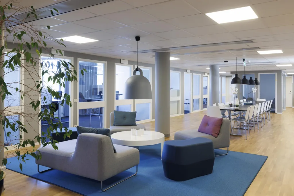 En lounge med soffor, bord, växter och moderna hängande lampor i ett rymligt kontor.