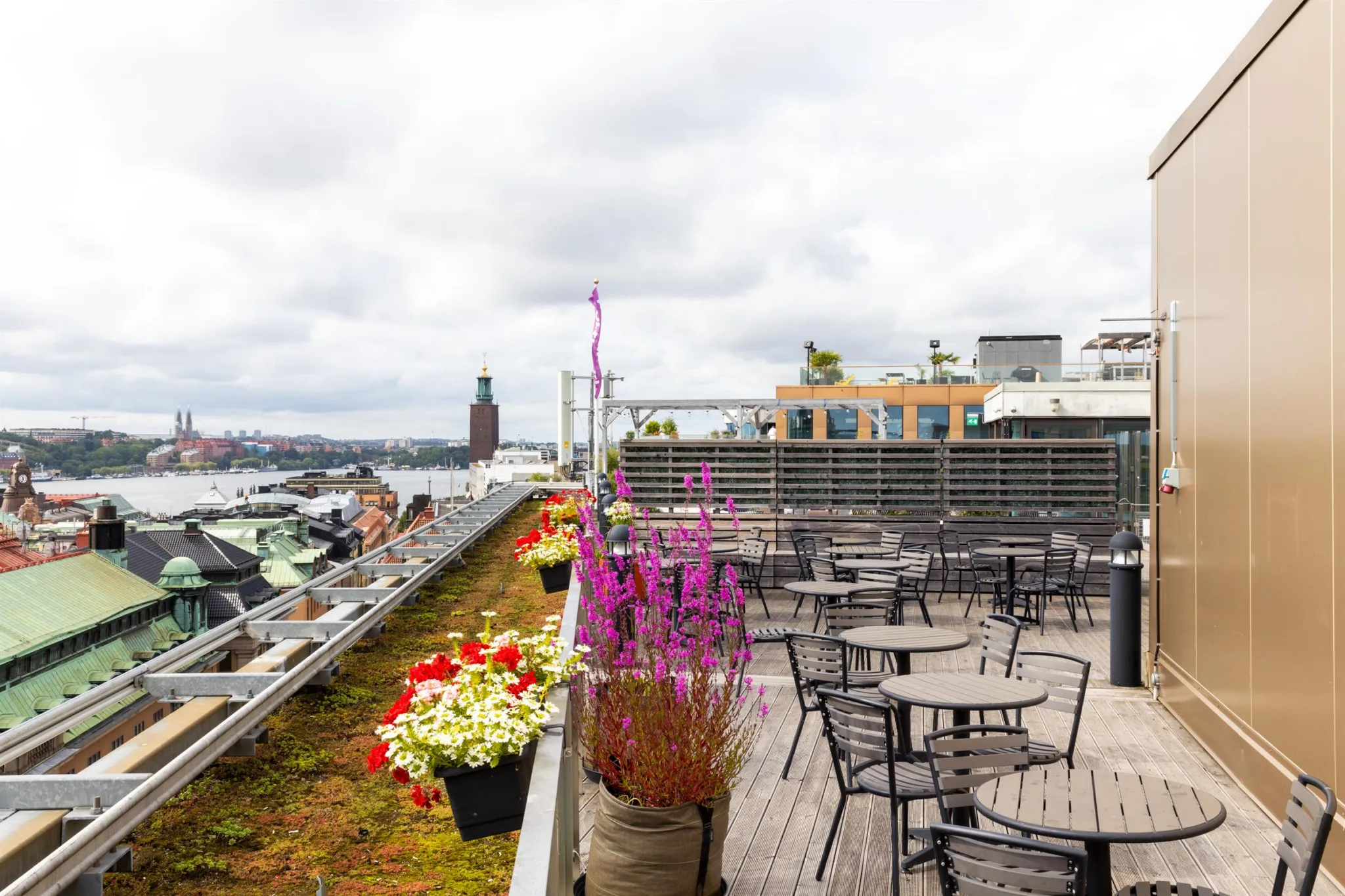 En takterrass med bord, stolar, färgglada blommor och utsikt över en stad med vatten och byggnader.
