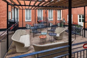En uteplats med rottingmöbler, små bord och trätrall, omgiven av röda tegelväggar.