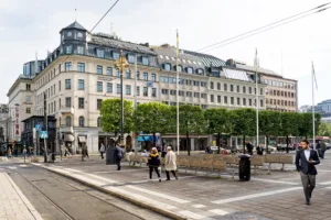 En stadsmiljö med en bred trottoar, människor som promenerar och en byggnad med klassisk arkitektur i bakgrunden.