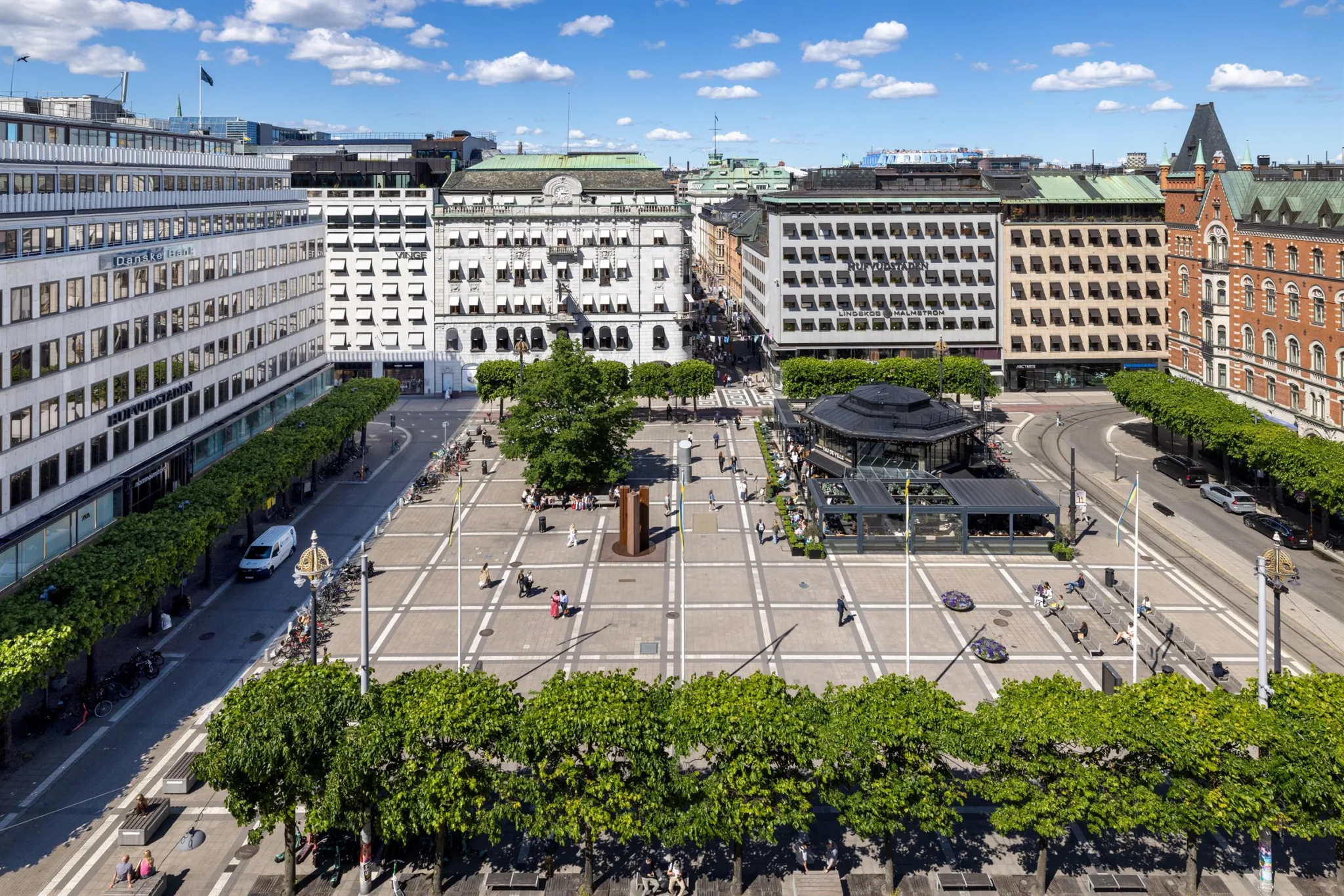 En överblick av ett torg med omgivande byggnader, träd och människor som rör sig på torget.