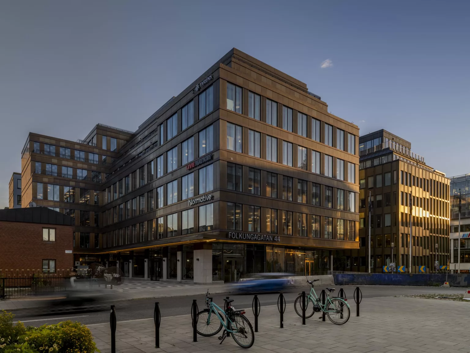 En modern byggnad med glasfasad i kvällsljus, cyklar parkerade framför och en bil på gatan.