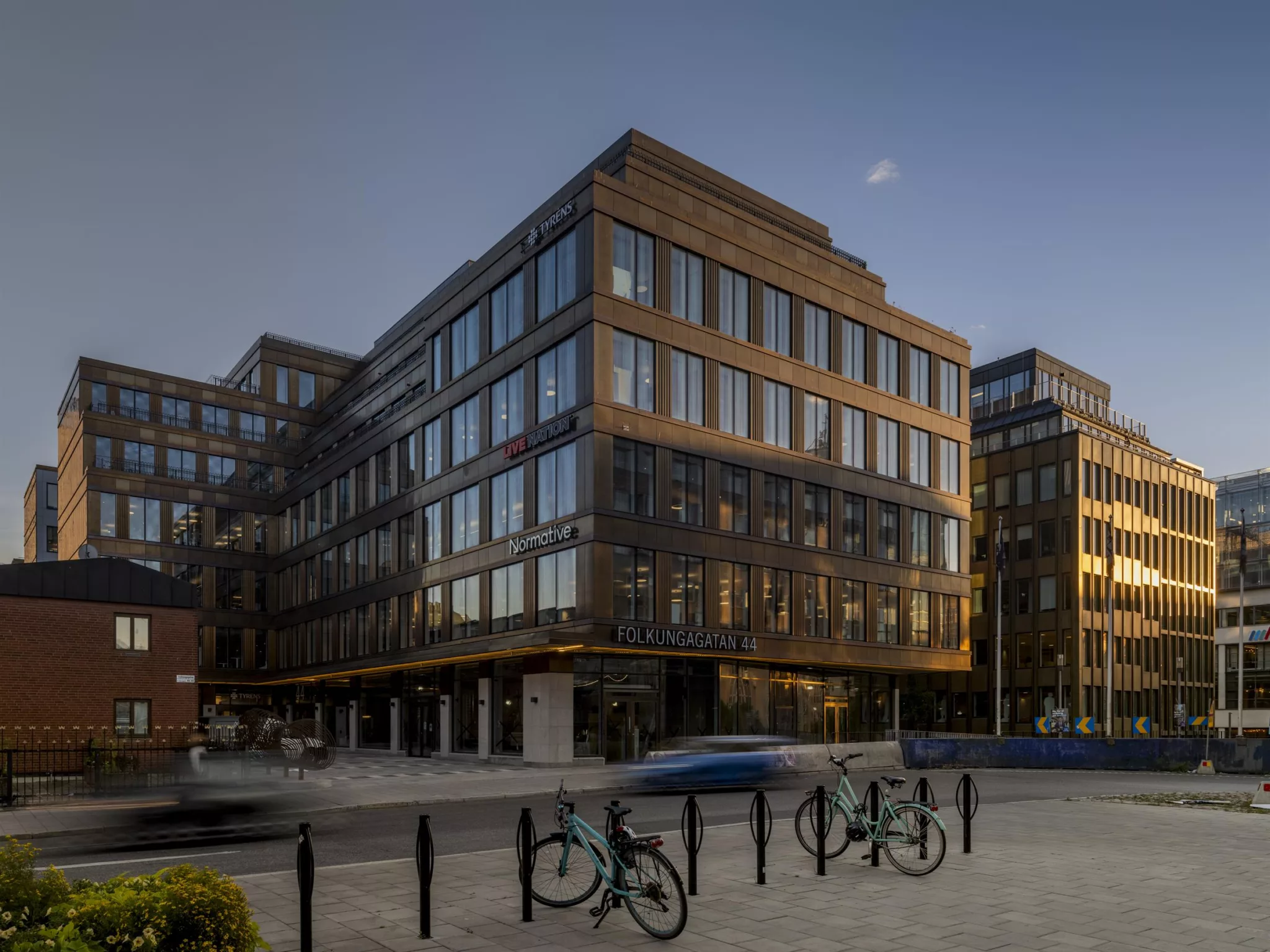 En modern byggnad med glasfasad i kvällsljus, cyklar parkerade framför och en bil på gatan.