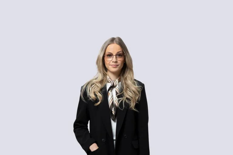 3. Kvinna med långt blont hår, glasögon, svart kavaj och vit scarf mot en ljusgrå bakgrund.