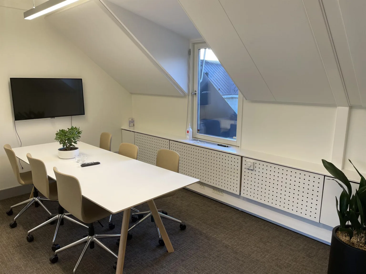 5. Ett mötesrum med vitt bord, beige stolar, växt och väggmonterad TV under ett snedtak.