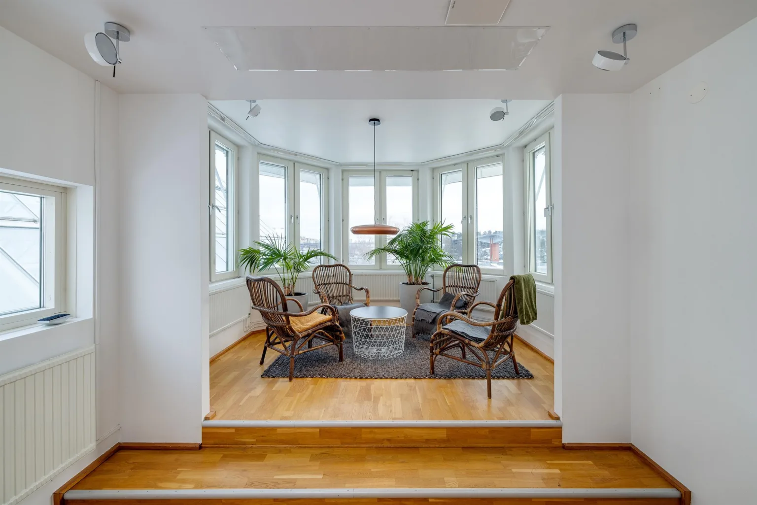 3. Litet rum med stora fönster, rottingmöbler, en matta, växter och en hängande lampa.