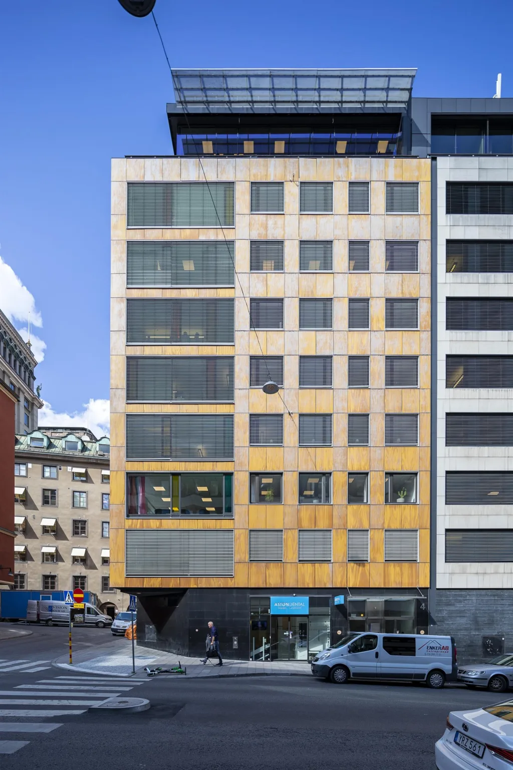 En modern byggnad med fasad i trä och glas, omgiven av andra byggnader och blå himmel.