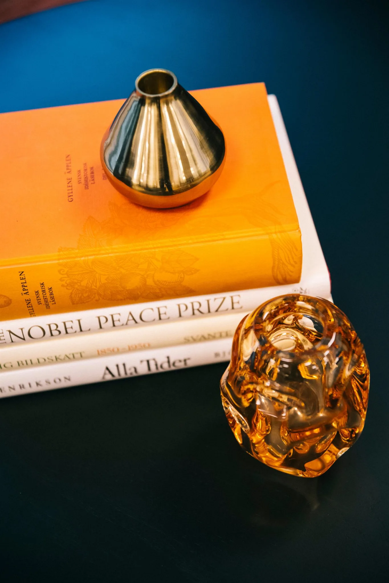 Två böcker i orange och vitt, en gyllene dekorativ figur och ett transparent ornament på ett mörkt bord.