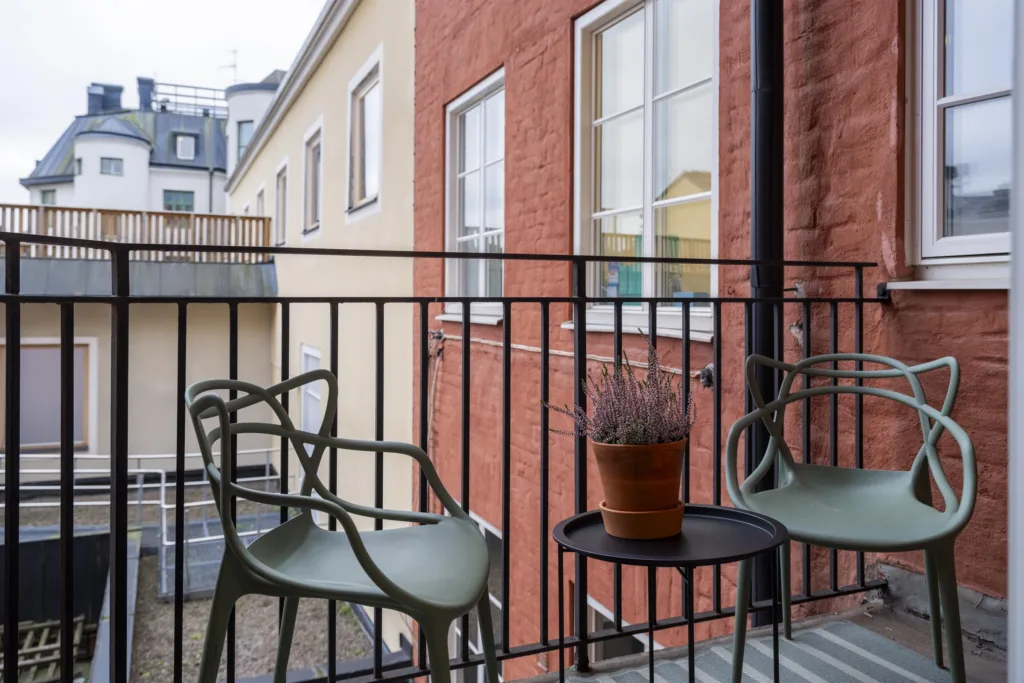 1. Balkong med två gröna stolar, ett litet bord och en krukväxt, omgiven av tegel- och putsade fasader.