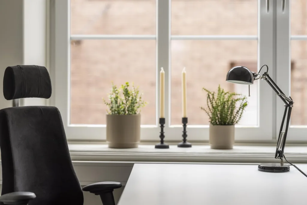 2. Ett skrivbord nära ett fönster med två krukväxter, två tända ljus och en skrivbordslampa.